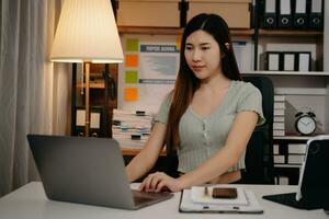 joven hermosa mujer trabajo tarde desde hogar en habitación a tarde noche horas extras.empresaria doblar terminado lugar de trabajo mientras mirando a ordenador portátil computadora. foto