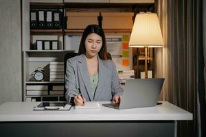 joven hermosa mujer trabajo tarde desde hogar en habitación a tarde noche horas extras.empresaria doblar terminado lugar de trabajo mientras mirando a ordenador portátil computadora. foto