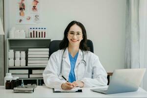 Confident young  Asian female doctor in white medical uniform sit at desk working on computer. Smiling use laptop write in medical journal photo