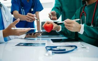 médico equipo teniendo un reunión con doctores en blanco laboratorio abrigos y quirúrgico matorrales sentado a un mesa que se discute un pacientes trabajando en línea utilizando ordenadores foto