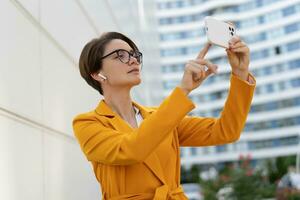 Smiling  beautiful  woman with short haircut  in yellow jacket and pants using mobyle phone. Standing over modern city. Outdoor photo. photo