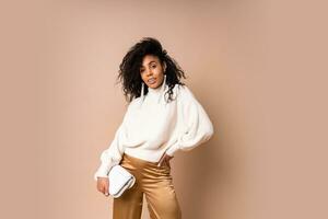 Fashion studio photo of elegant  african american woman in casual white sweater and silk pants posing on beige background. Looking at camera.