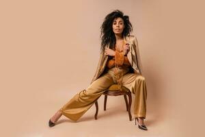 Beautiful  african model with perfect curly hairs in elegant orange blouse and silk pants sitting on vintage chair onver beige background. photo