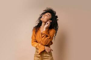 Glad african woman with perfect curly hairs in casual orange blouse and golden pants posing on beige background. photo