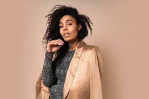 Confident  black woman in golden silk jaket and shiny sexy dress with perfect tan body posing in studio over beige background. photo