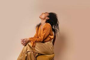 Laughing  african model with perfect curly hairs in elegant orange blouse and silk pants sitting on vintage chair onver beige background. photo