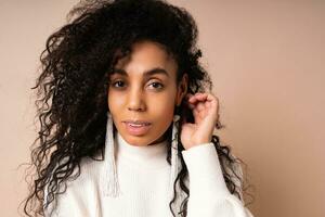 Fashion portrait of elegant mix race woman with curly hairs in white casual  sweater posing in studio on beige background. photo