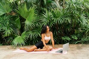 Graceful woman in stylish sport outfit  looking online training and  sitting on yoga matt over tropical garden. Bodycare and welness concept. photo