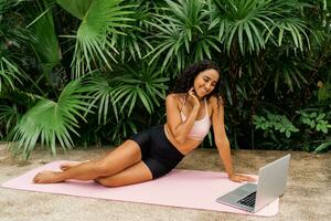 Graceful woman in stylish sport outfit  looking online training and  sitting on yoga matt over tropical garden. Bodycare and welness concept. photo