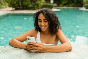 Smiling woman with curly hairstyle making self portrait by mobylephone while chilling in swimming pool. photo