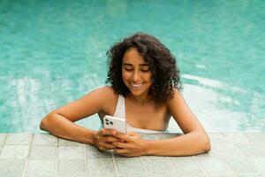 Smiling woman with curly hairstyle making self portrait by mobylephone while chilling in swimming pool. photo