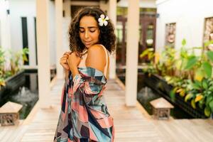 Outdoor summer photo of pretty brunette woman with tropical flower in hairs posing in  stylish  hotel.