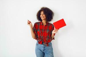 Pensive African student female with book posing  indoor over white  wall. Wearing red checkered shirt. Blue jeans. photo