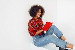 elegante americano mujer con Rizado pelos sentado en clase habitación y escritura en cuaderno. foto