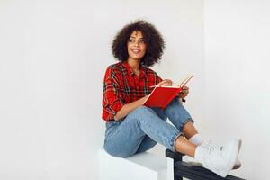 Stylish American woman with curly  hairs sitting  in class room and writing in notebook. photo