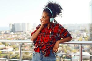 Enthusiastic American woman with dark skin posing with earphones on the rooftop. Urban landscape background. photo