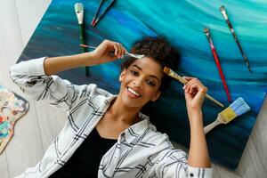 Top view of happy  painter   woman lying on canvas   and looking at the camera with  brushes  in hands. photo