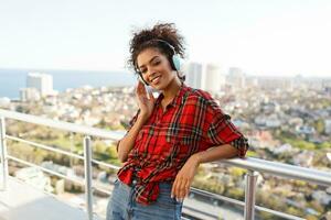 Close up portrait of  American woman enjoying lovely music by earphones  , dressed in checkered shirt, standing on rooftop. Urban landscape background. photo