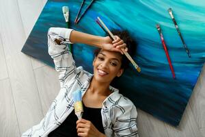 Top view of happy  painter   woman lying on canvas   and looking at the camera with  brushes  in hands. photo