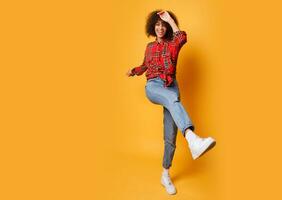 Active  girl with stylish Afro hairs  dancing on bright orange background. Studio photo of happy curly lady in  red shirt jumping and  having fun indoor. Full length.