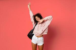 Portrait of  confident sexy black woman in stylish hoodie   with back pack posing on pink background. photo