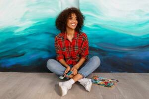 Beautiful  cute african ethnicity student  holding brushes and posing over her abstract artworks on canvas. photo