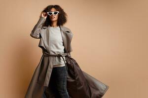 Black woman in trendy grey leather jacket posing over beige background in studio. Winter and autumn fashion look. photo