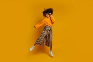 Cute African girl in orange sweater and colorful pants  jumping and funny dancing on yellow background. Full length. photo