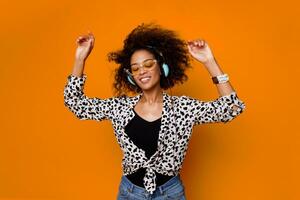African woman dancing and listening music by earphones in studio over orange background. Party mood. Optimistic mood. photo