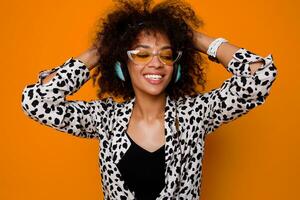 Studio portrait of  laughing  cute mix race woman with dark skin listening music by stylish earphones over orange background. Positive party mood. Afro hairs. photo