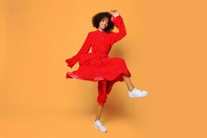 Dreamy mood. Stylish African girl dancing and jumping over orange background. Wearing red vintage dress . Shopping and fashion. photo