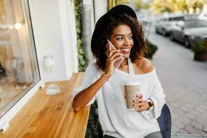 Traveling mix race  woman in stylish casual outfit relaxing outdoor  in city cafe , drinking coffee and chatting by mobile phone. Wearing trendy accessories and sunglasses. Spring mood. photo