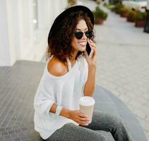 Close up portrait of Successful black woman, blogger or store manager  talking by mobile phone during coffee break. Sitting on stairs and holding paper cup of hot beverage. photo