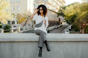 Outdoor positive image of smiling pretty black woman in white sweater and black hat  enjoying   coffee to go.  Urban background. photo