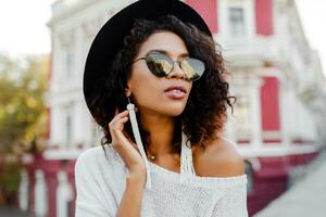 Close up portrait of fashionable black woman with stylish Afro hairs posing outdoor. Urban background. Wearing black sunglasses, hat and white earrings. Trendy accessories. Perfect smile. photo