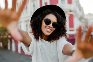 Beautiful black woman with stylish Afro hairs making self portrait. Wearing sunglasses, black hat and elegant earrings. Happy emotions. American city background. Travel girl. photo