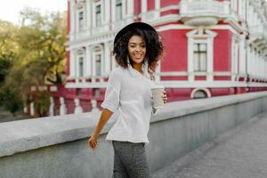 Outdoor positive image of smiling pretty black woman in white sweater and black hat holding cup of coffee.  Urban background. photo