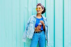 Happy mixed race female with stylish Afro hairs posing outdoor , holding cherry lemonade , wearing jeans jacket and blue wool sweater. photo