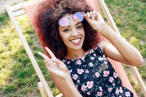 Happy fashionable black woman  with stylish curly hairstyle sitting on chaise-lounge on amazing green lawn in summer park with her friends on picnic. photo