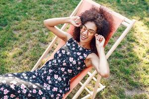 Happy fashionable black woman  with stylish curly hairstyle sitting on chaise-lounge on amazing green lawn in summer park with her friends on picnic. photo