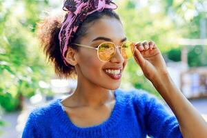 Close up portrait of  smiling black woman in trendy glasses. Bright pink bandana on hairs. Green trees on background. photo