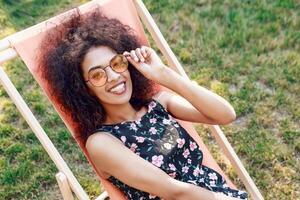 Happy fashionable black woman  with stylish curly hairstyle sitting on chaise-lounge on amazing green lawn in summer park with her friends on picnic. photo