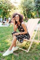 Happy fashionable black woman  with stylish curly hairstyle sitting on chaise-lounge on amazing green lawn in summer park with her friends on picnic. photo