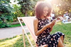 Mixed race girl sitting on chaise-lounge on lawn and  texting message on mobile phone. Summer park on background .  Pretty woman spending her  weekend with friends on nature. photo