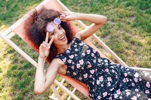 Happy young mixed rave woman with amazing curly hairs relaxing on chaise-longue  on green lawn in park . Wearing stylish summer dress . photo