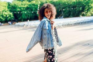 juguetón mujer envuelve sí misma terminado su hombro y mirando en cámara. agotador. elegante hipster atuendo. afro peinado . Perfecto verano ocio hora en el noche parque. foto