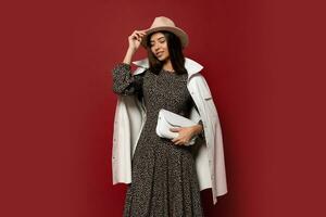 Autumn fashion look. Studio image of gorgeous european brunette girl in trendy white jacket and dress with print posing on red background. Holding leather handbag. photo