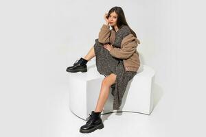 Graceful european woman in winter fur coat and stylish dress  sitting in studio over white background. Wearing  ankle boot in black leather. photo