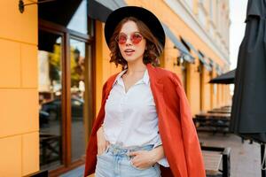 Outdoor fashion image of young elegant woman in orange jacket and white blouse walking in sunny city. photo