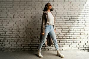 Stylish short haired woman in casual leather coat and jeans  posing over urban brick wall. Street style look. Autumn mood. Full lenght. photo